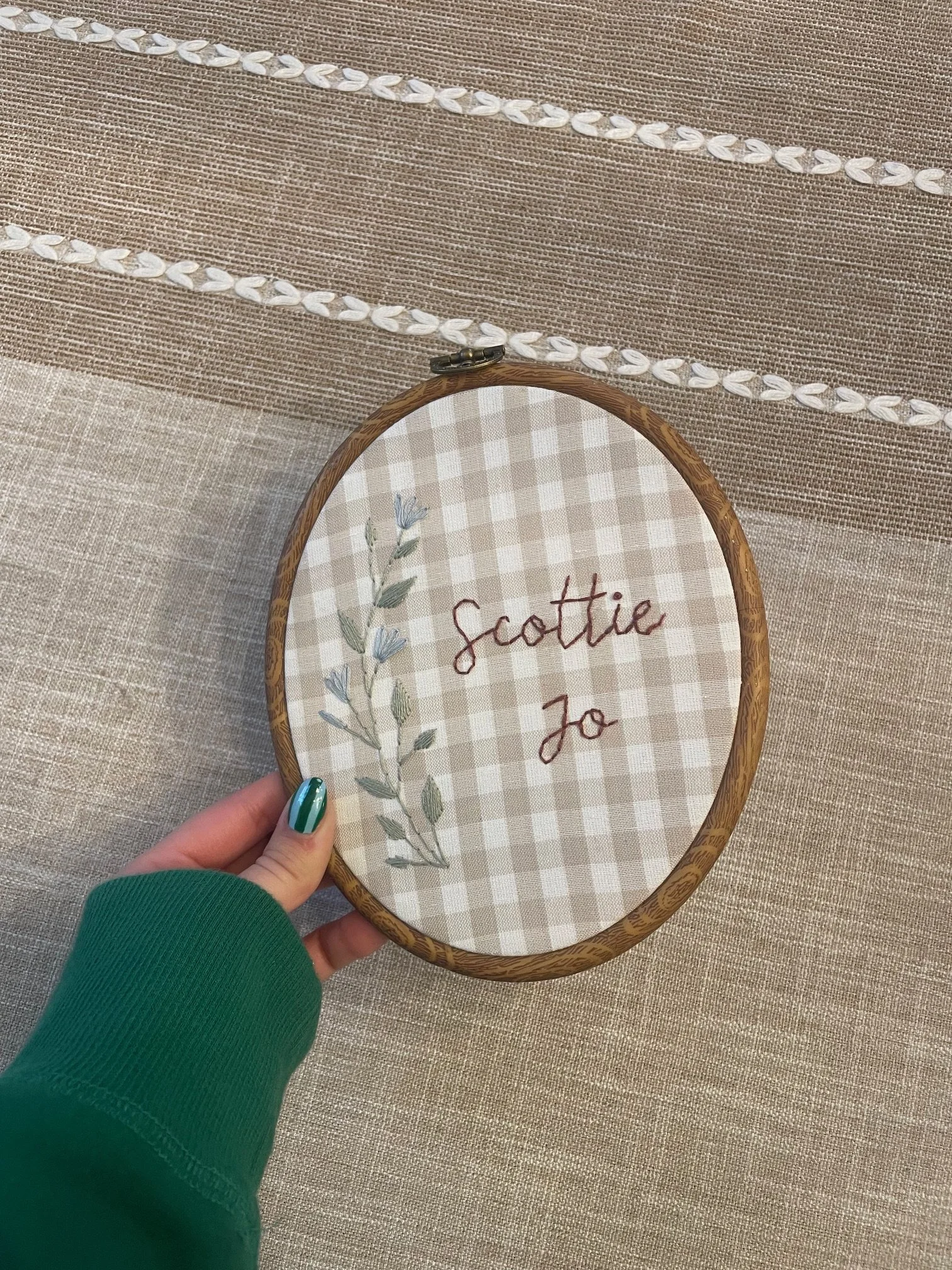 Hand holding an embroidered embroidery hoop with the words "Scottie Jo" on beige checkered fabric, on a textured beige tablecloth.