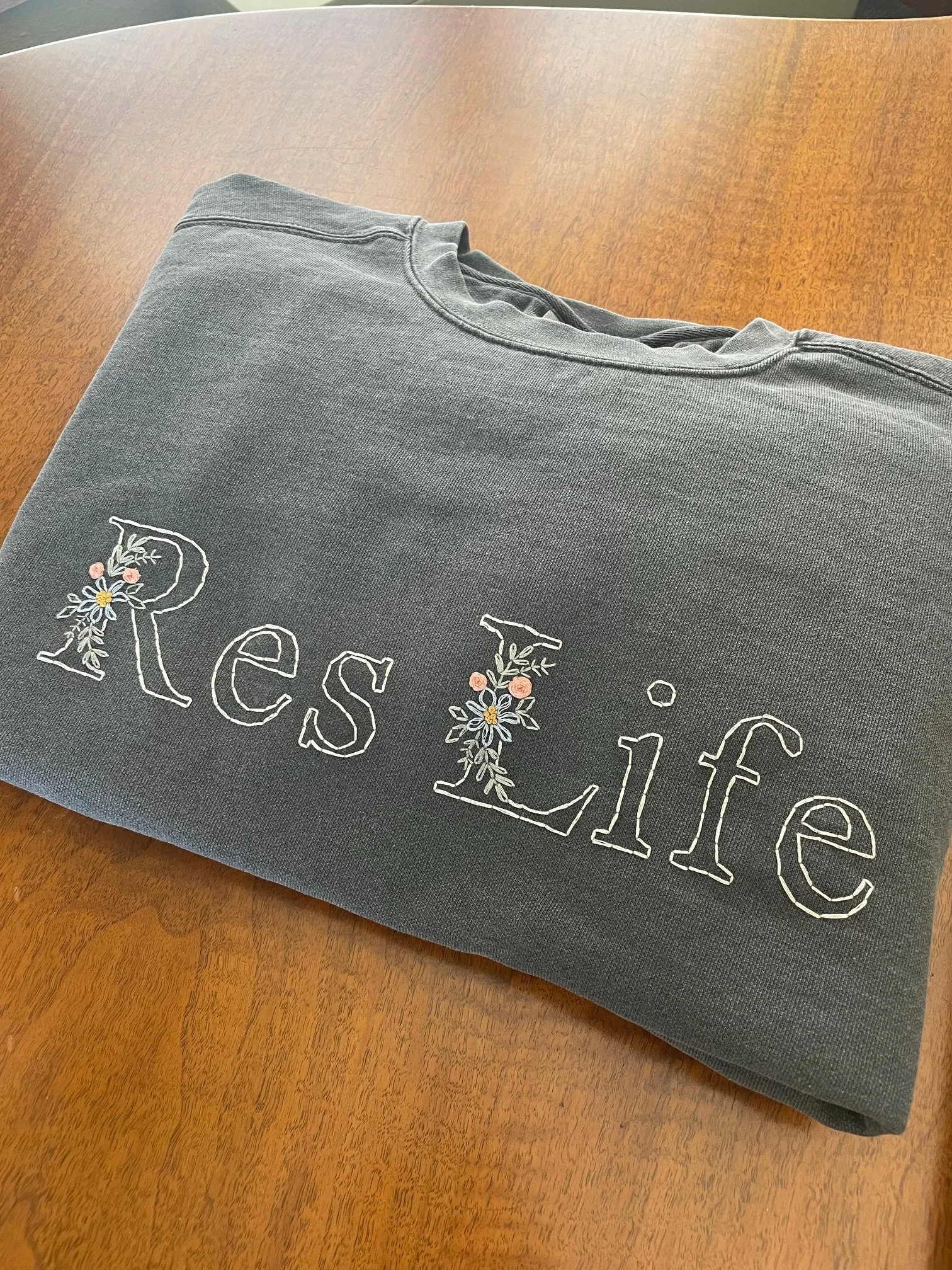Gray sweatshirt with embroidered floral design and partial text, placed on a wooden table.
