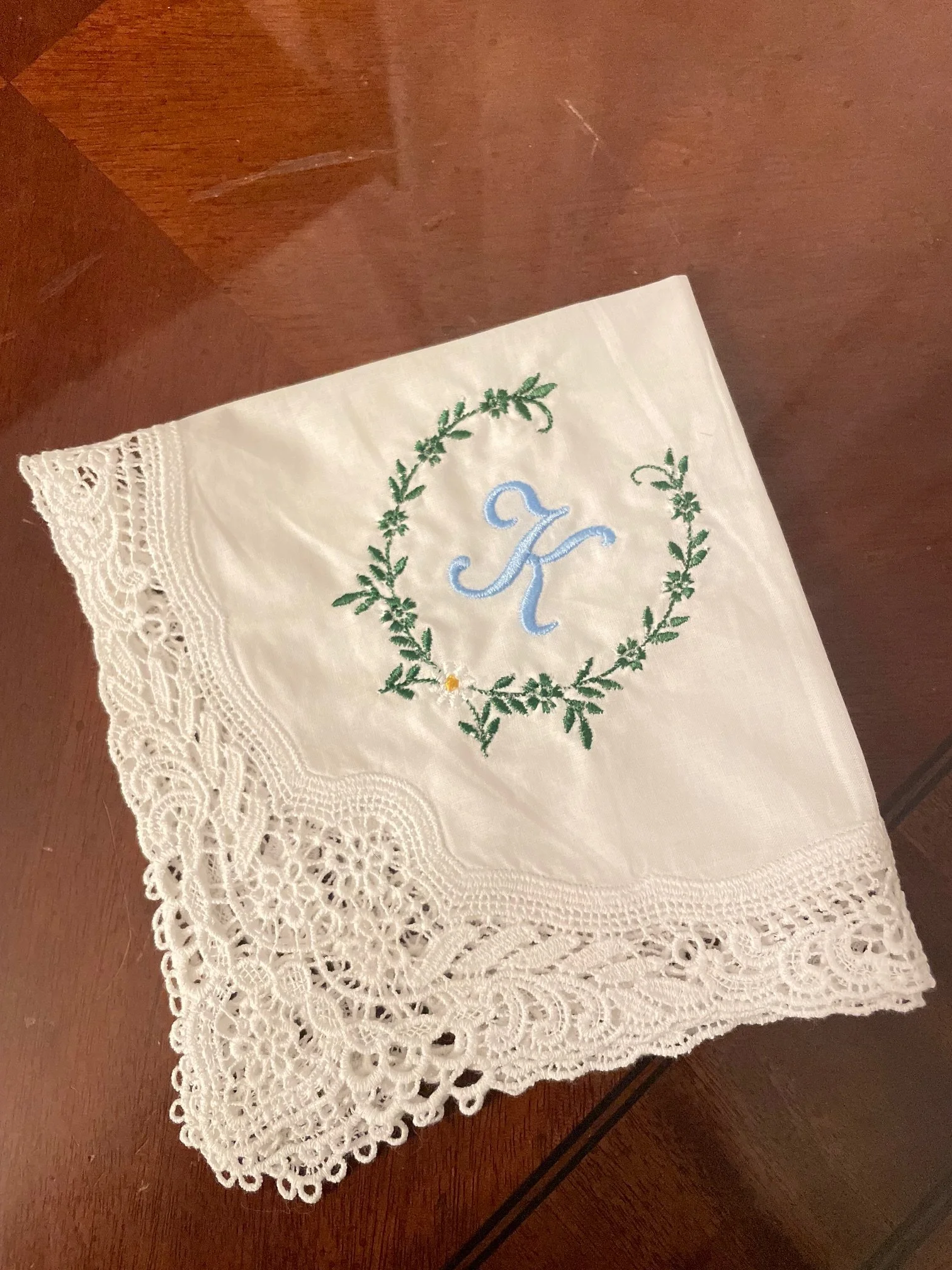 A white cloth napkin with embroidered green leaves, a blue letter 'K' in the center, and intricate white lace trim on a wooden surface.