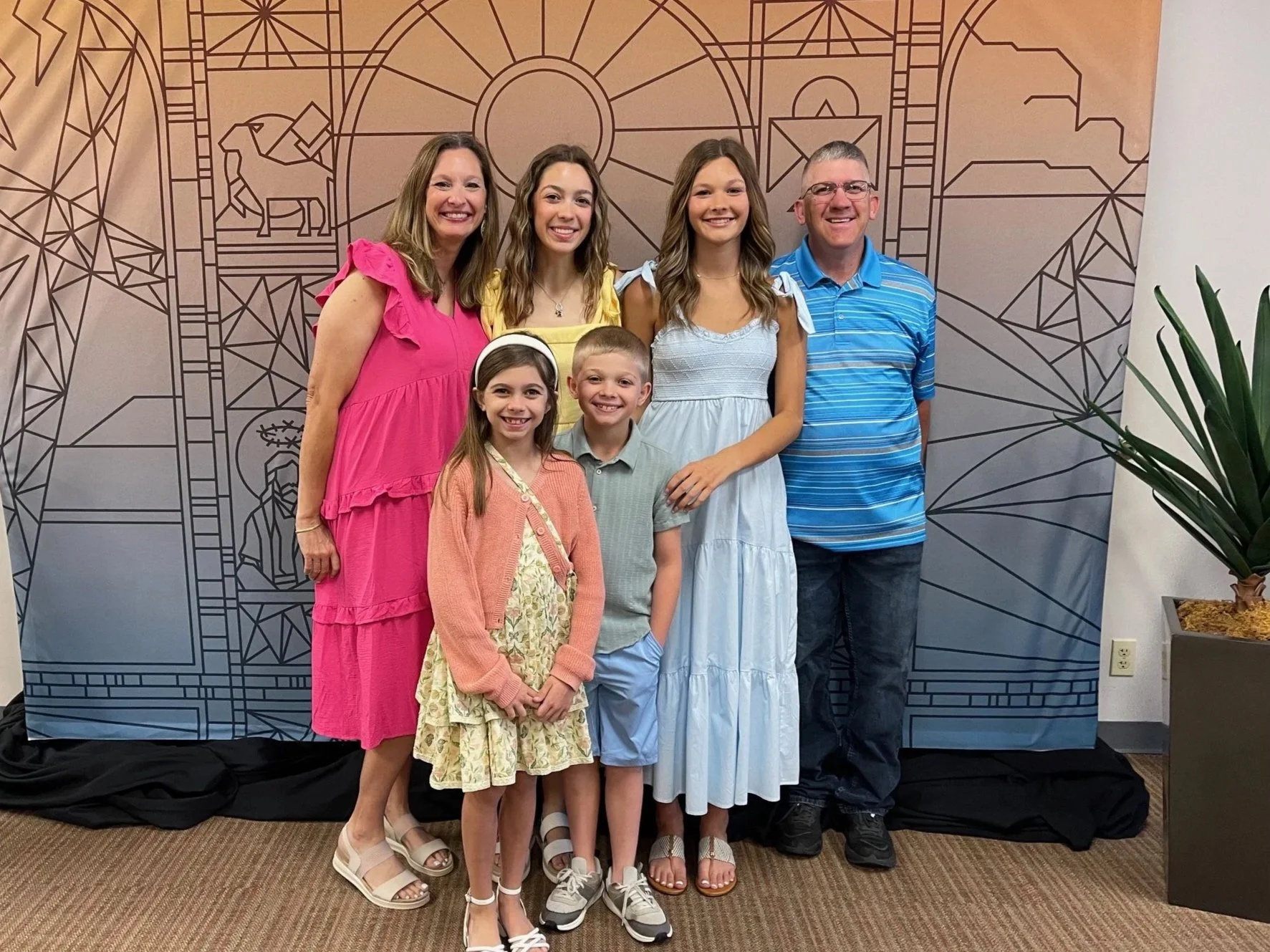 A group of seven smiling people, including three children and four adults, posing together in front of a geometric backdrop with religious symbols such as a lion and a circle. They are indoors, standing on a carpeted floor, with a potted plant visible on the right.