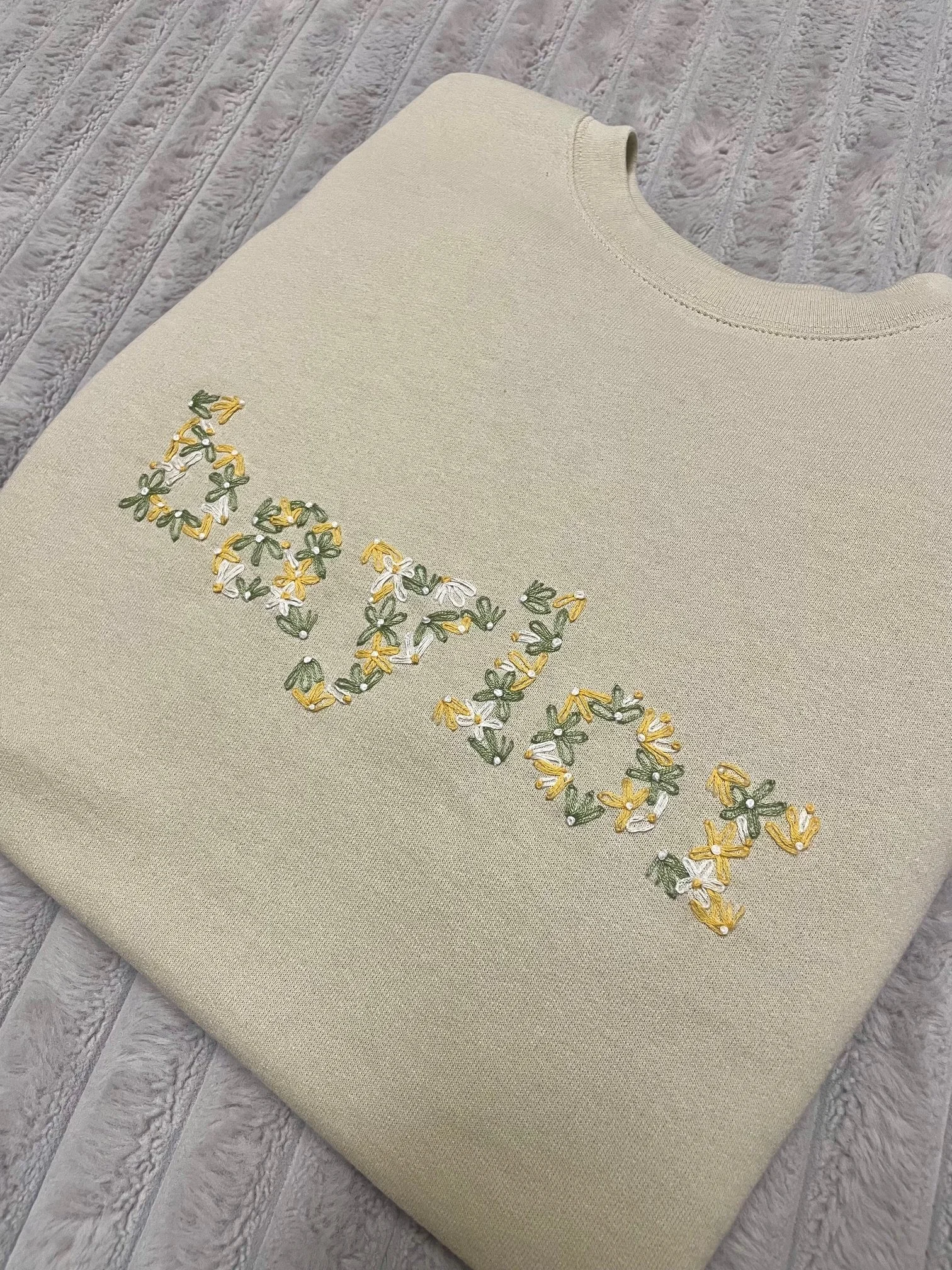 Close-up of a beige sweater with embroidered yellow, white, and green flowers and the word 'love' on top of a textured wooden surface.