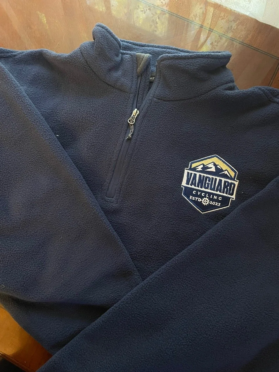 A navy blue fleece jacket with a zipped collar and an embroidered logo that says "Vanguard Cycling est. 2023" on the left chest.