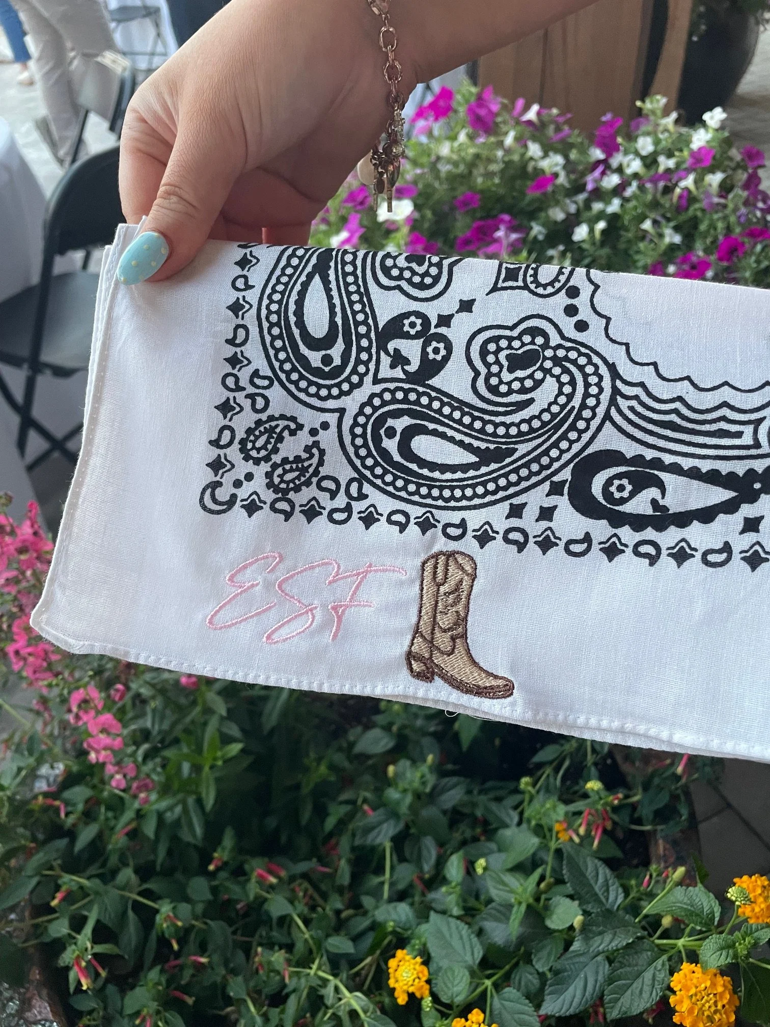 A hand holding a white bandana with black paisley patterns and pink embroidery, with a background of pink, purple, and yellow flowers.