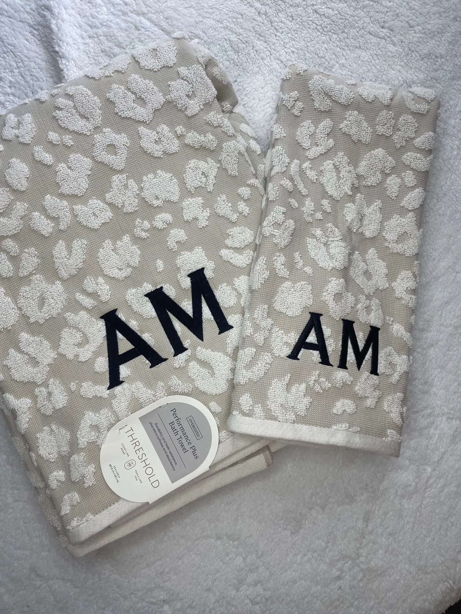 A folded bath towel with a white textured floral pattern, featuring large dark blue letters partially visible, on a soft, grey surface.