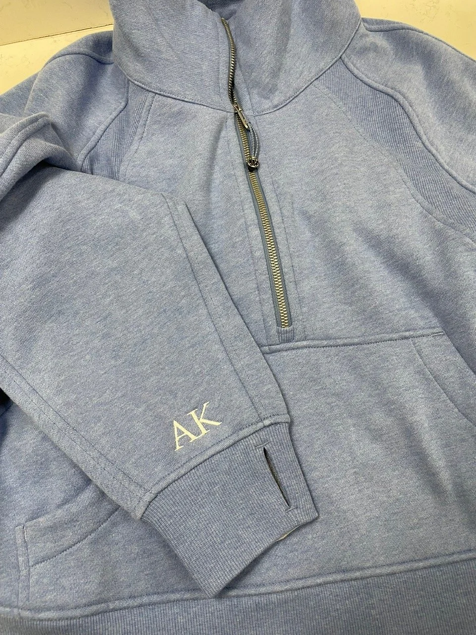 A light gray quarter-zip sweatshirt with embroidered initials 'AK' on the sleeve and a zipper pocket on the chest.