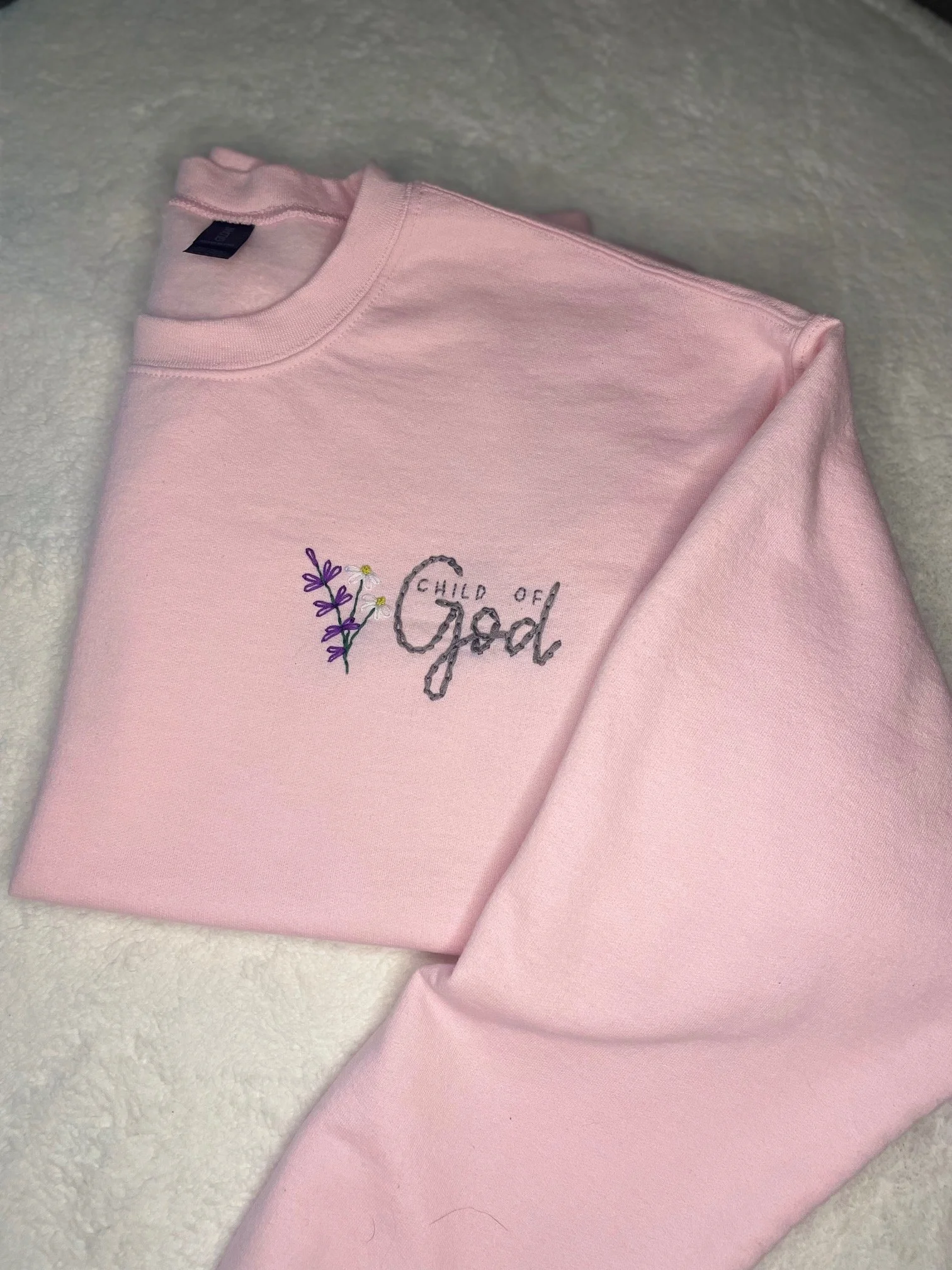 A light pink sweatshirt with floral embroidery and text on the front, folded and placed on a soft, textured white surface.