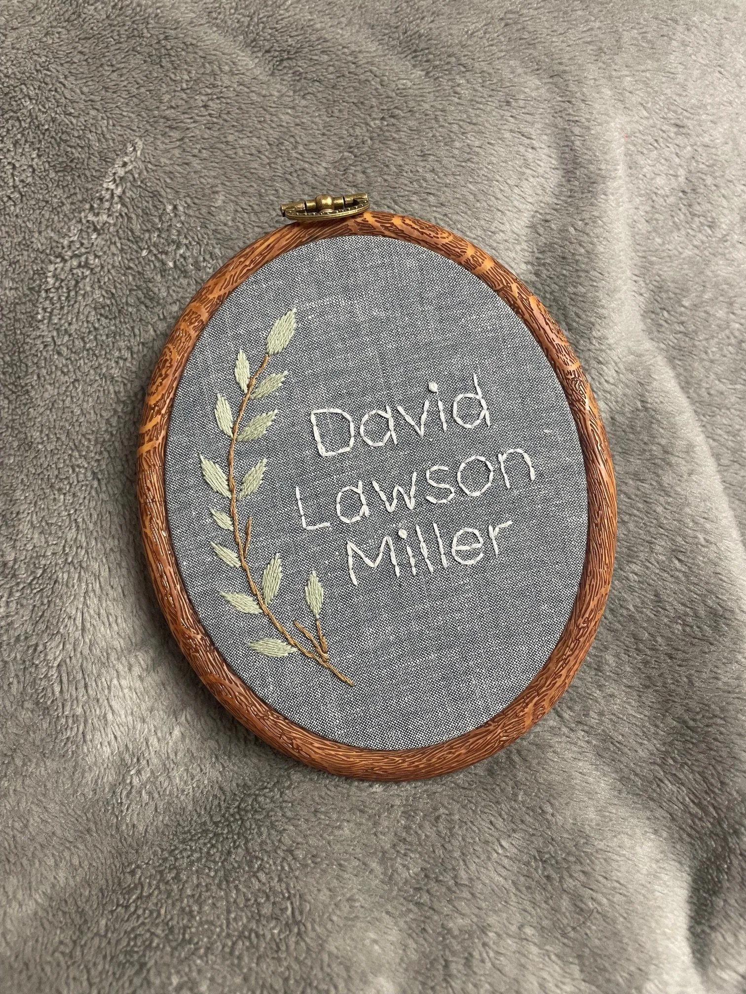 An embroidered oval fabric piece with a wooden embroidery hoop, displaying hand-stitched text that reads 'David Lawson Miller' and a leaf branch design, placed on a soft gray textured surface.