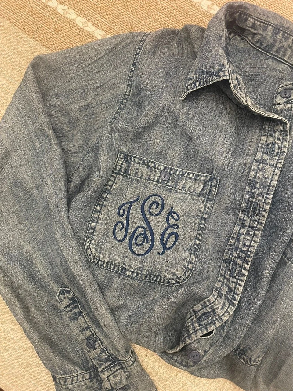 A gray, long-sleeved, button-up denim shirt with a monogrammed pocket that has the initials 'JSG' embroidered in navy thread.