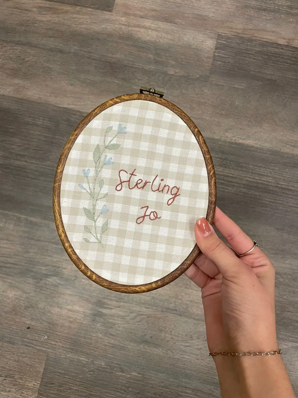 Image shows a wooden embroidery hoop with a beige gingham fabric in it. It shows the name "sterling jo" and a floral branch hand stitched into the fabric.