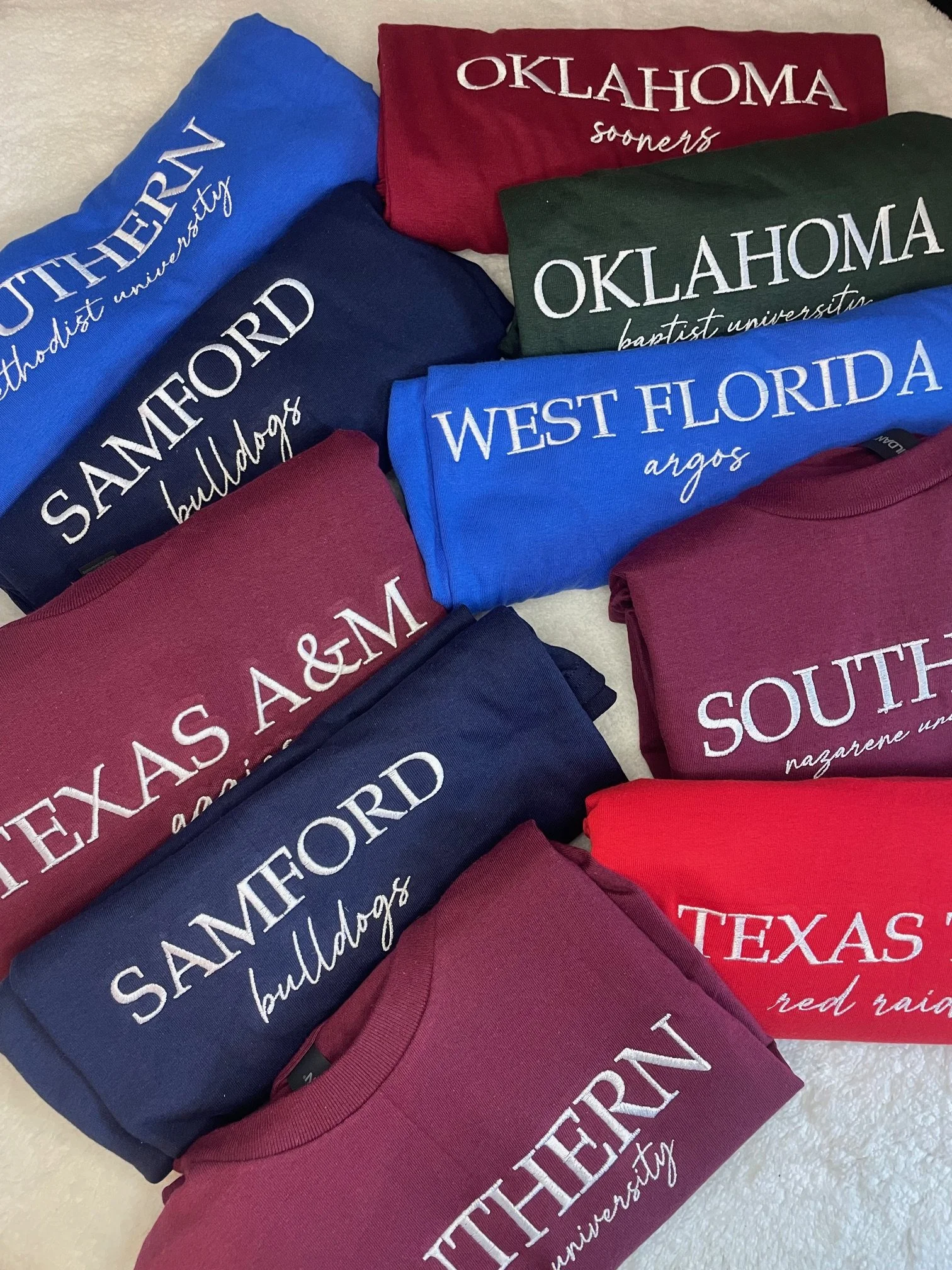Stacks of folded T-shirts in various colors, each with text referencing Oklahoma cities and universities, including Oklahoma Sooners, Samford Bulldogs, West Florida Argos, South Texas, and Texas A&M.