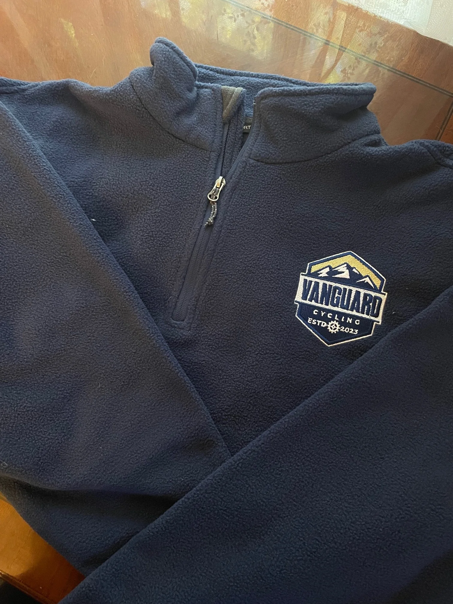 A navy blue fleece jacket with a half-zip, standing collar, and a logo patch on the left side that reads "Vanguard Cycling Est. 2023" along with a mountain graphic.