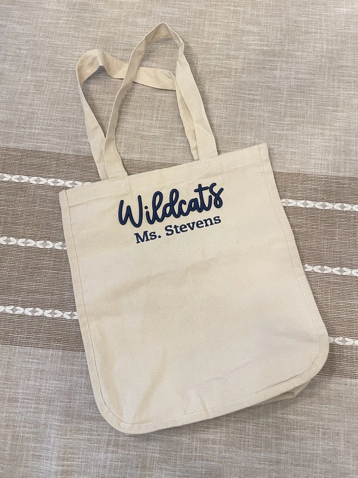 A beige tote bag with navy text that reads 'Wildcats Ms. Stevens' resting on a beige fabric surface with white decorative embroidery.