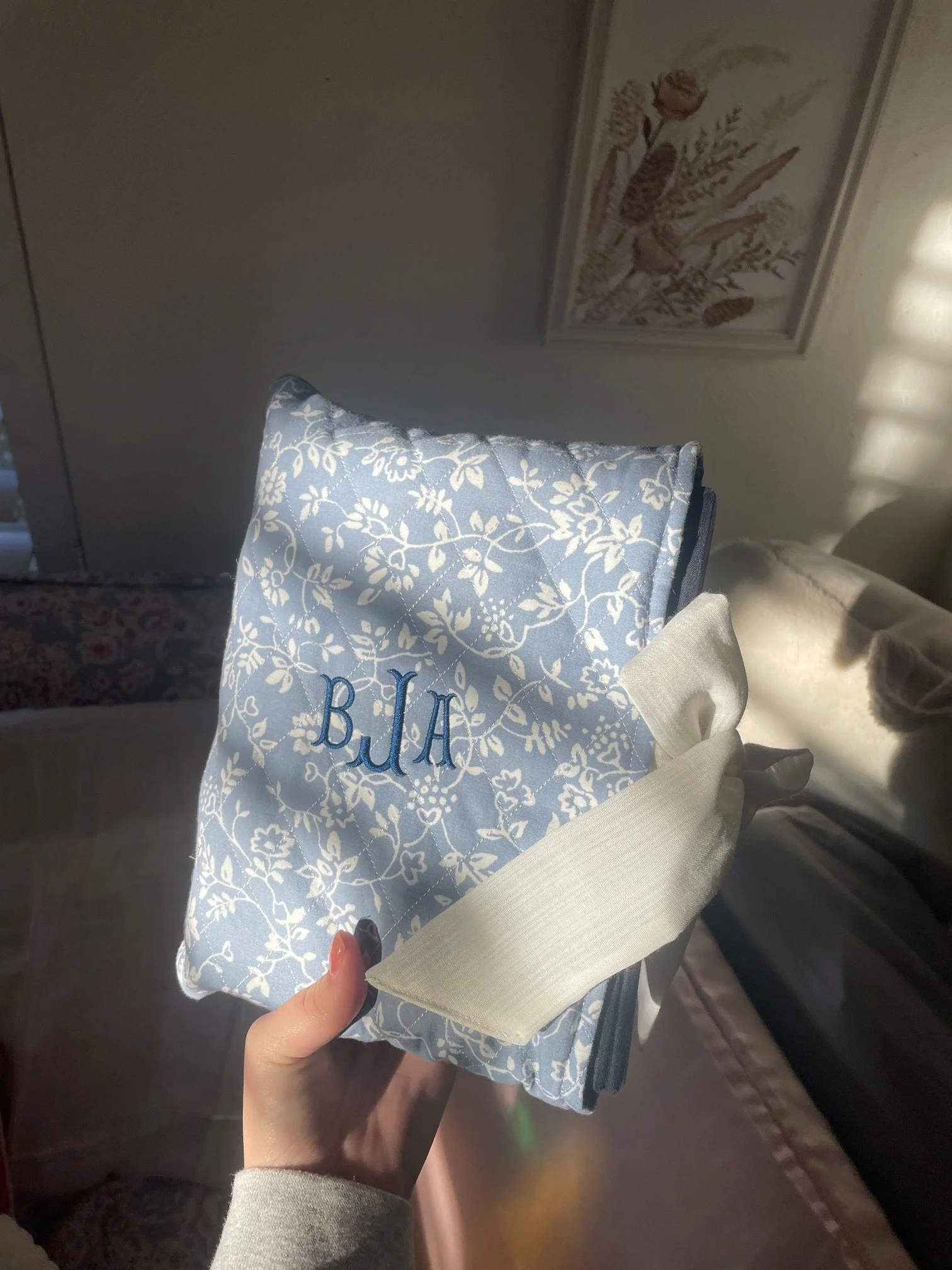 Person holding a small gift bag with blue and white floral design and the word "BLAH" embroidered on it, in an indoor setting with artwork on the wall.