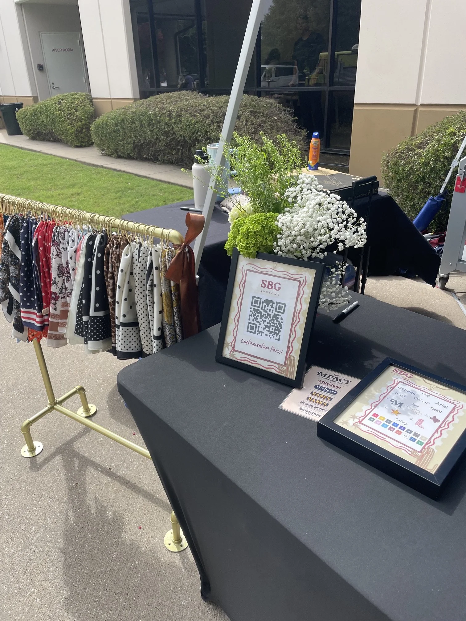 Outdoor booth display with clothing on a gold rack, potted plants, and sign with QR code for a shop called SBC Custom. The booth is set up on a sidewalk next to a building with bushes and reflective windows.
