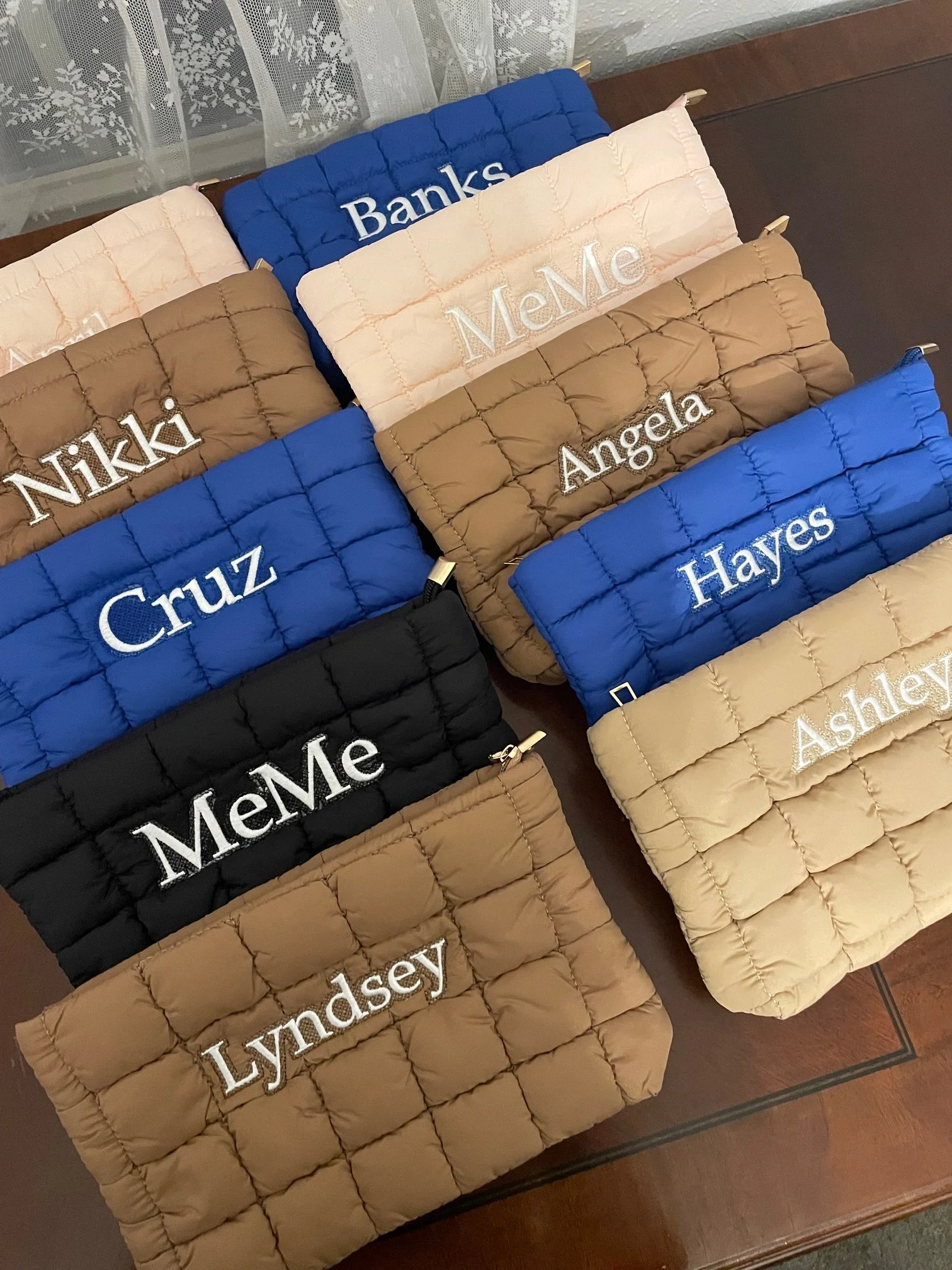 A display of quilted fabric pouches with embroidered names in white thread, arranged on a wooden surface. The pouches come in various colors, including black, tan, navy blue, and blush pink, with each name such as Nikkie, Cruz, Angeles, MeMe, Lyndsey