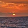 Sunset over the ocean with an orange sun near the horizon and calm water in the foreground.