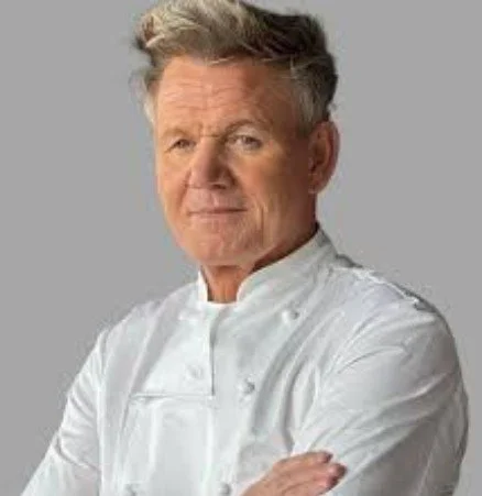 A senior male chef with gray hair wearing a white chef's coat, standing with arms crossed against a grey background.