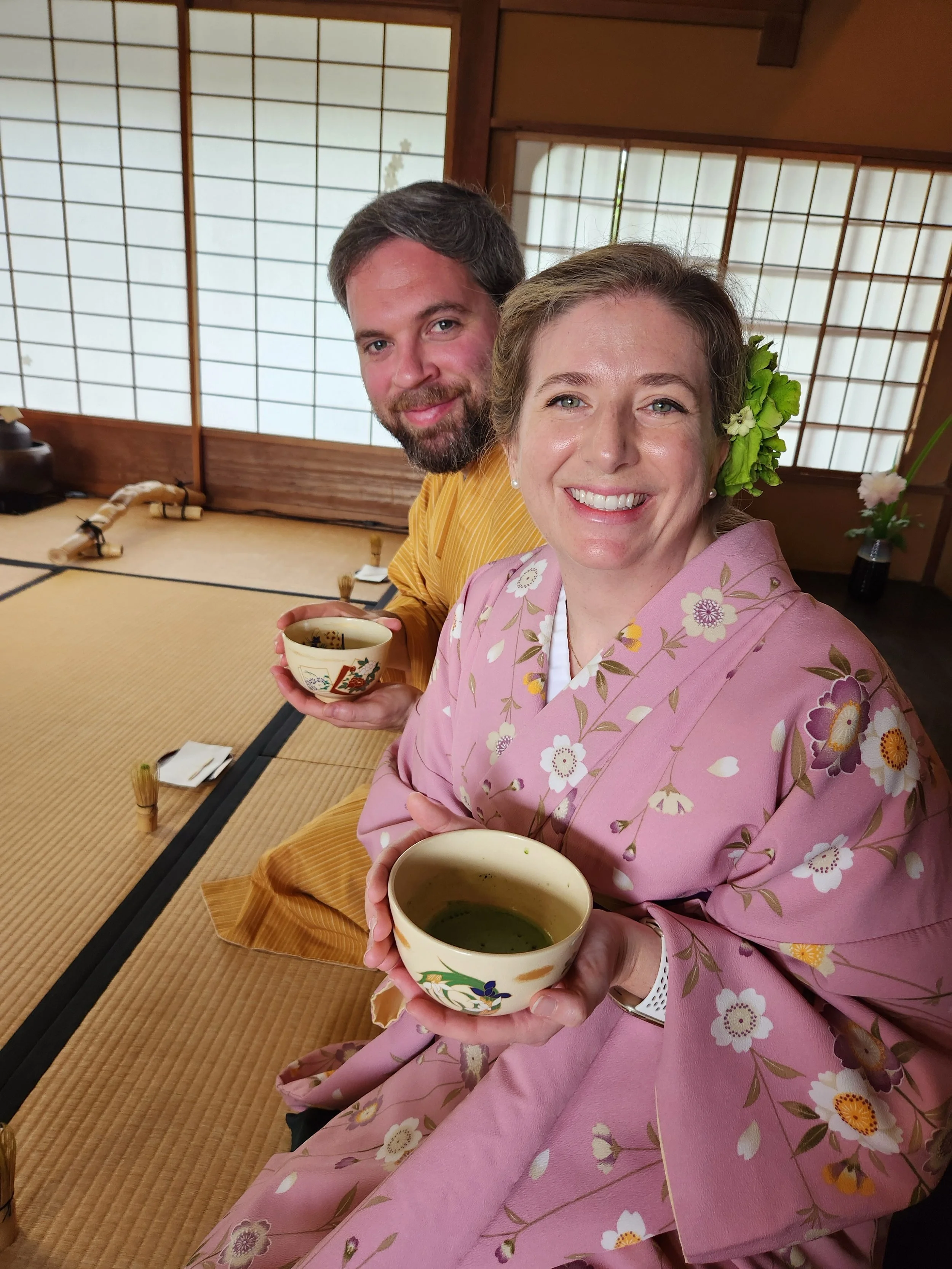 Two people dressed in traditional Japanese kimonos sitting on tatami mats in a room with shoji screens, holding bowls of green tea, smiling at the camera.