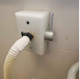 A white wall-mounted device with a hose, a dial, and a black circular indicator, used for industrial or maintenance purposes.