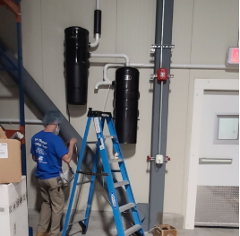 Technician working on pipes and equipment on a wall inside an industrial or building facility, with a blue ladder and various control valves.