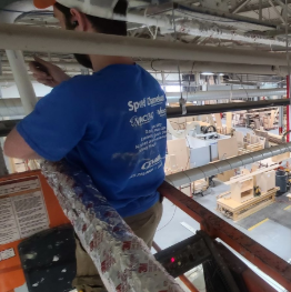 A worker wearing a blue shirt working on scaffolding inside a tall industrial or construction site, with machinery and other construction materials visible in the background.