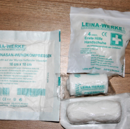First aid supplies including a sealed package of large bandages, a small package of first aid wipes, and a wrapped ice pack on a wooden surface.