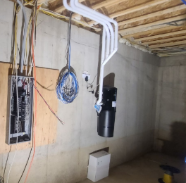 Electrical panel with wires, conduit, and a black sump pump or similar device installed on the wall in an unfinished basement.