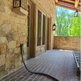 Back porch with brick flooring, stone wall, and glass door, with a black hose on the ground and a view of green trees in the background.
