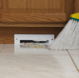 A broom sweeping up debris into a floor vent in a tiled kitchen floor.