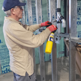 A man wearing glasses, a cap, beige shirt, and red gloves installing or working on a metal wall frame with a cordless drill and a yellow spray can in a construction site.