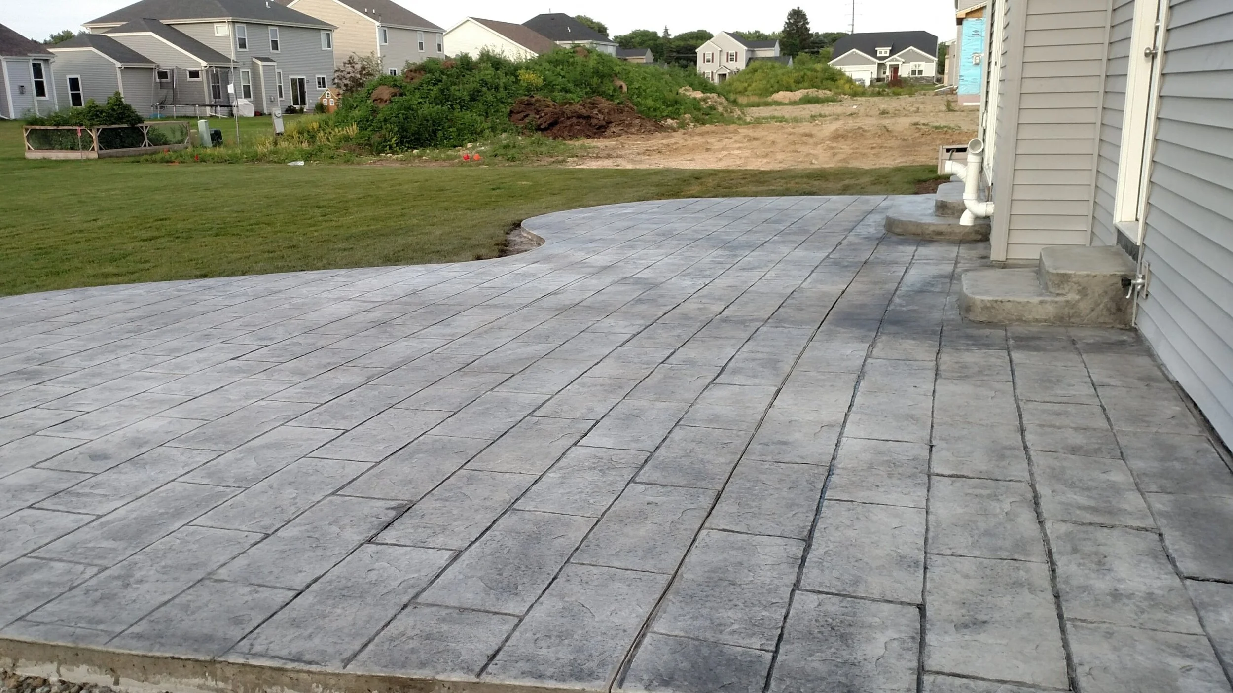 Newly constructed gray concrete patio with paving stones in a suburban backyard, with grass, dirt, and houses in the background.