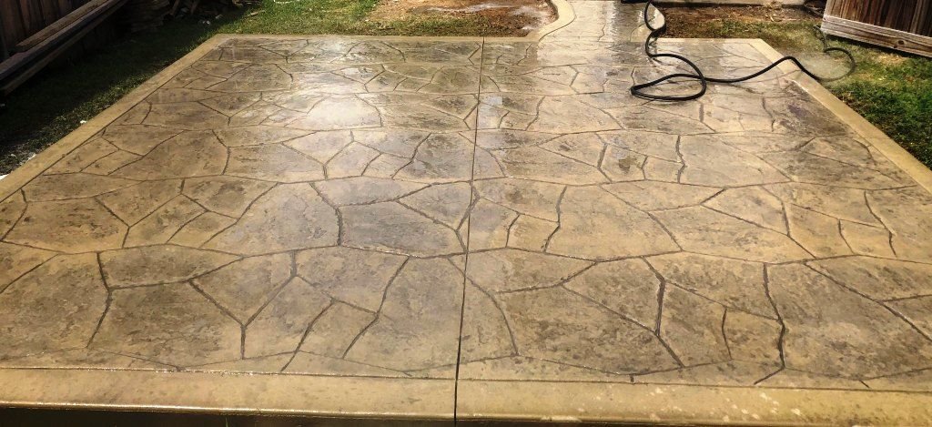A newly stamped concrete patio with a stone pattern in a backyard, partially wet from recent washing or sealing, with a garden hose on the right side.