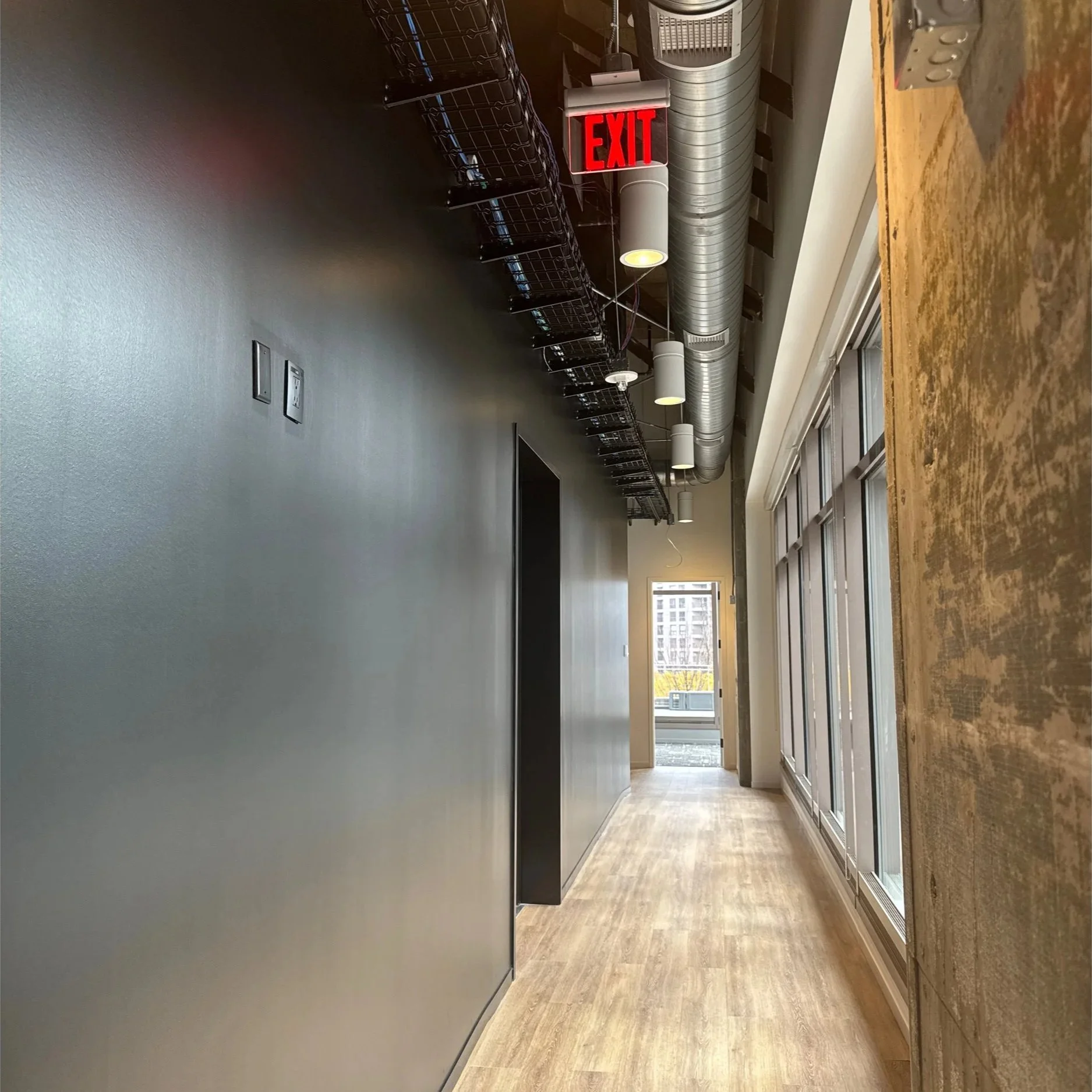 Atlanta Journal-Constitution Studio - Interior view of a modern office or commercial space with a hallway, metal storage racks, a red illuminated exit sign, ventilation ducts, and large windows at the end of the corridor.