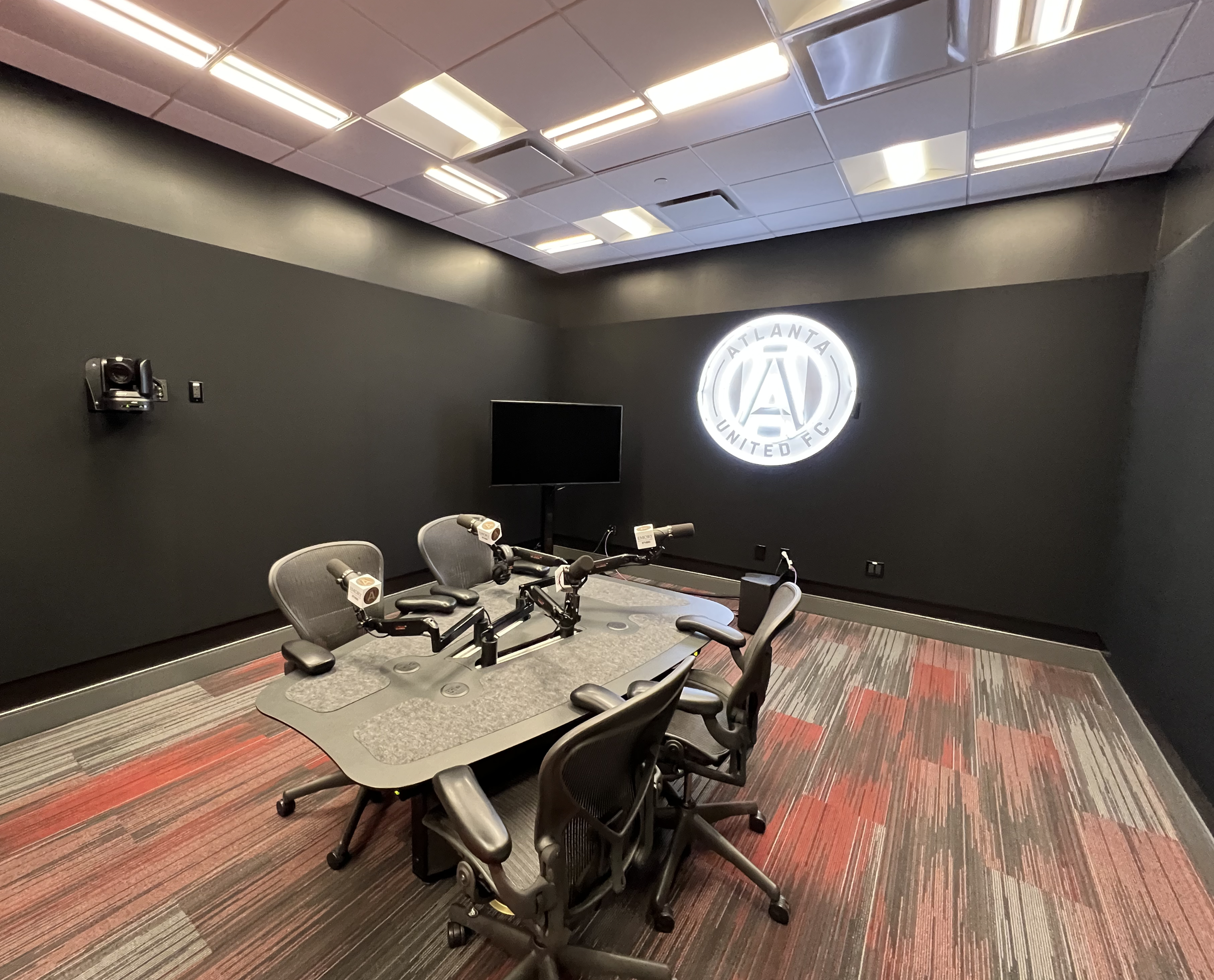 Conference room with black walls, a large illuminated Atlanta United FC logo, chairs around a table, microphones, a television screen, and ceiling lights.