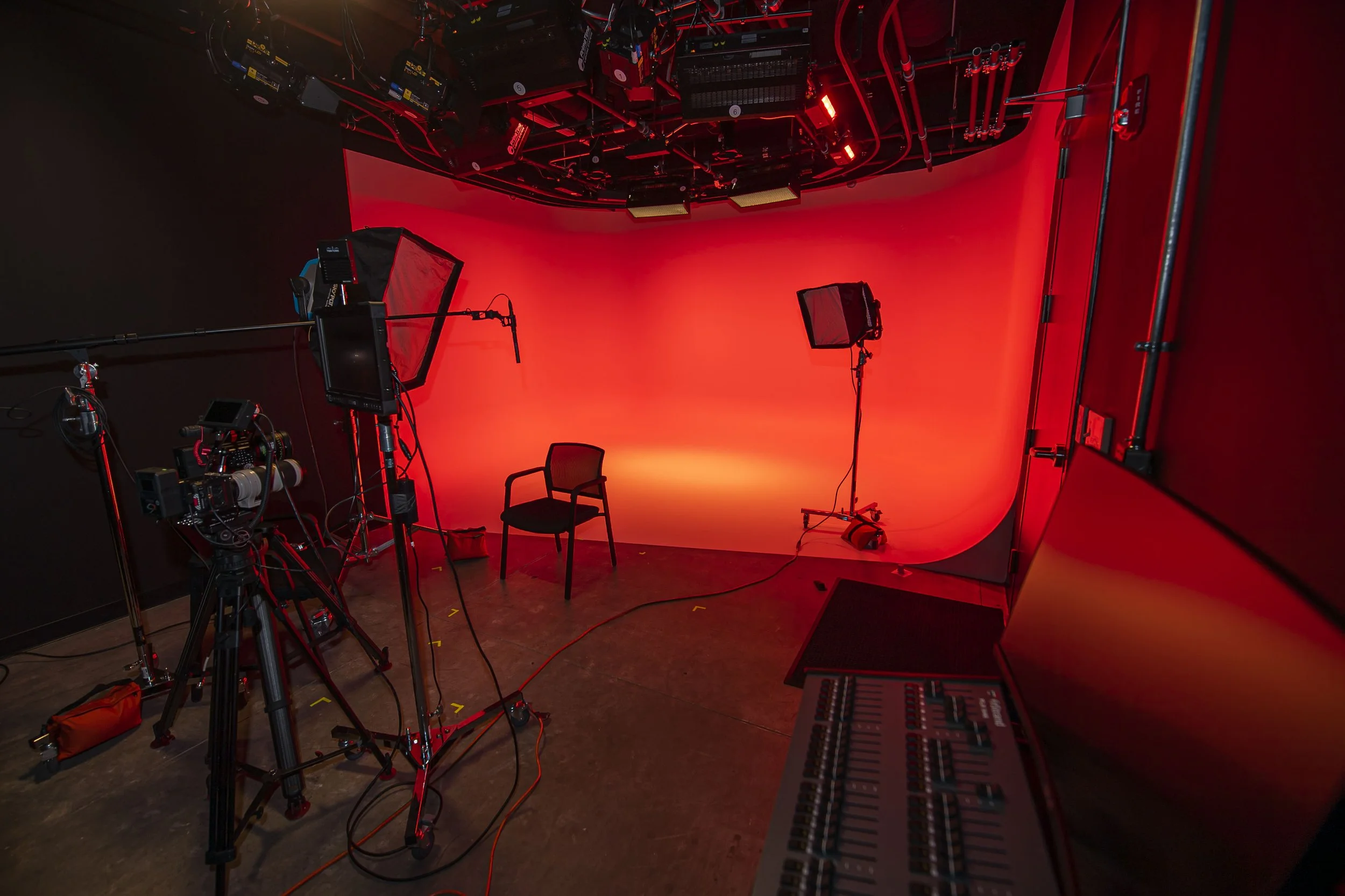 A professional photography studio setup with red background walls, two studio lights on stands directed towards the background, a black chair centered in front of the background, and filming equipment including cameras and a camera crane on the left side. Cables and a mixing console are also visible.