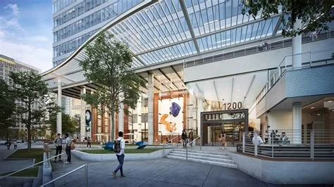 Modern office building entrance with glass facade, surrounded by people walking, trees, and outdoor seating.