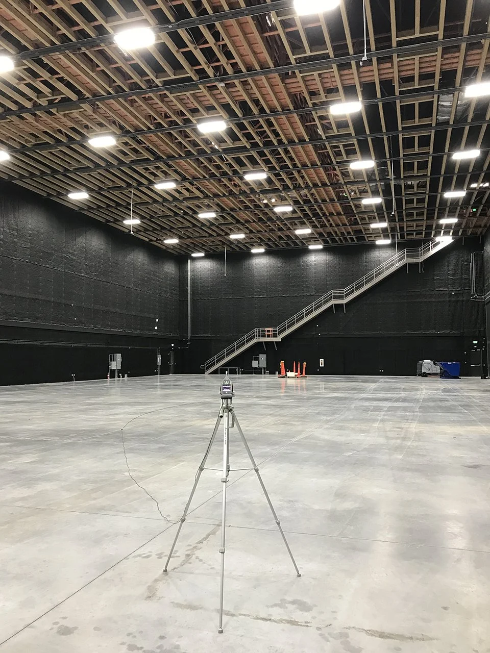 Empty industrial or warehouse space with high ceiling, lighting, and a tripod with a scanner or camera in the foreground.