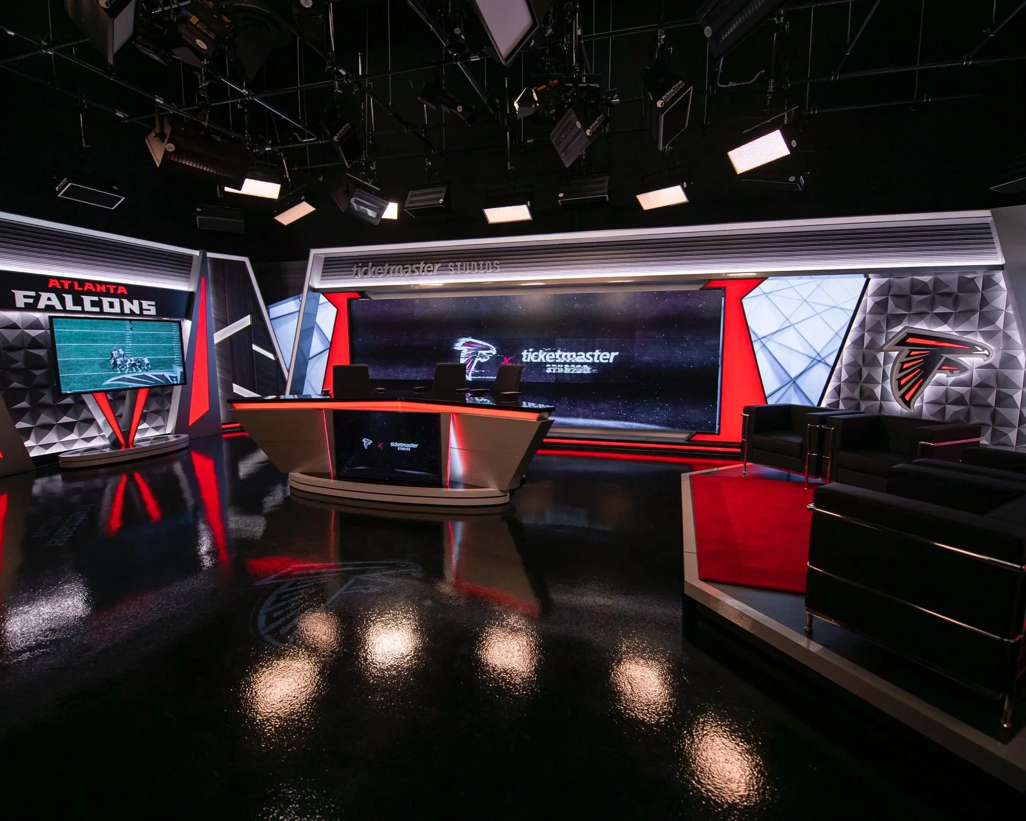 Studio with black shiny floor, black walls, illuminated signs for Atlanta Falcons and ticketmaster, large screen, black chairs, modern design with geometric patterns, overhead lighting fixtures.