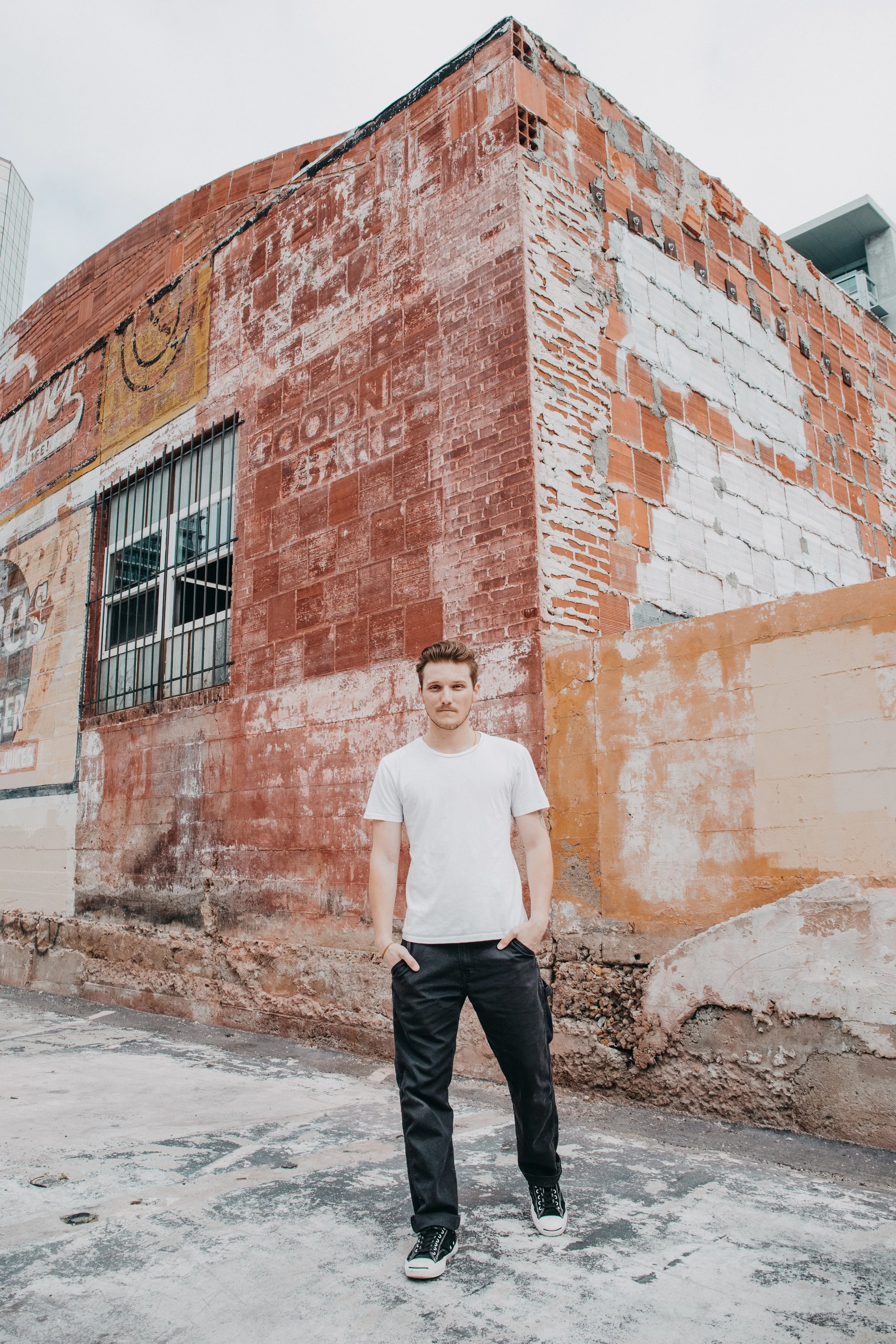 Young man in white t-shirt and black jeans standing in front of a weathered brick wall on an urban street.
