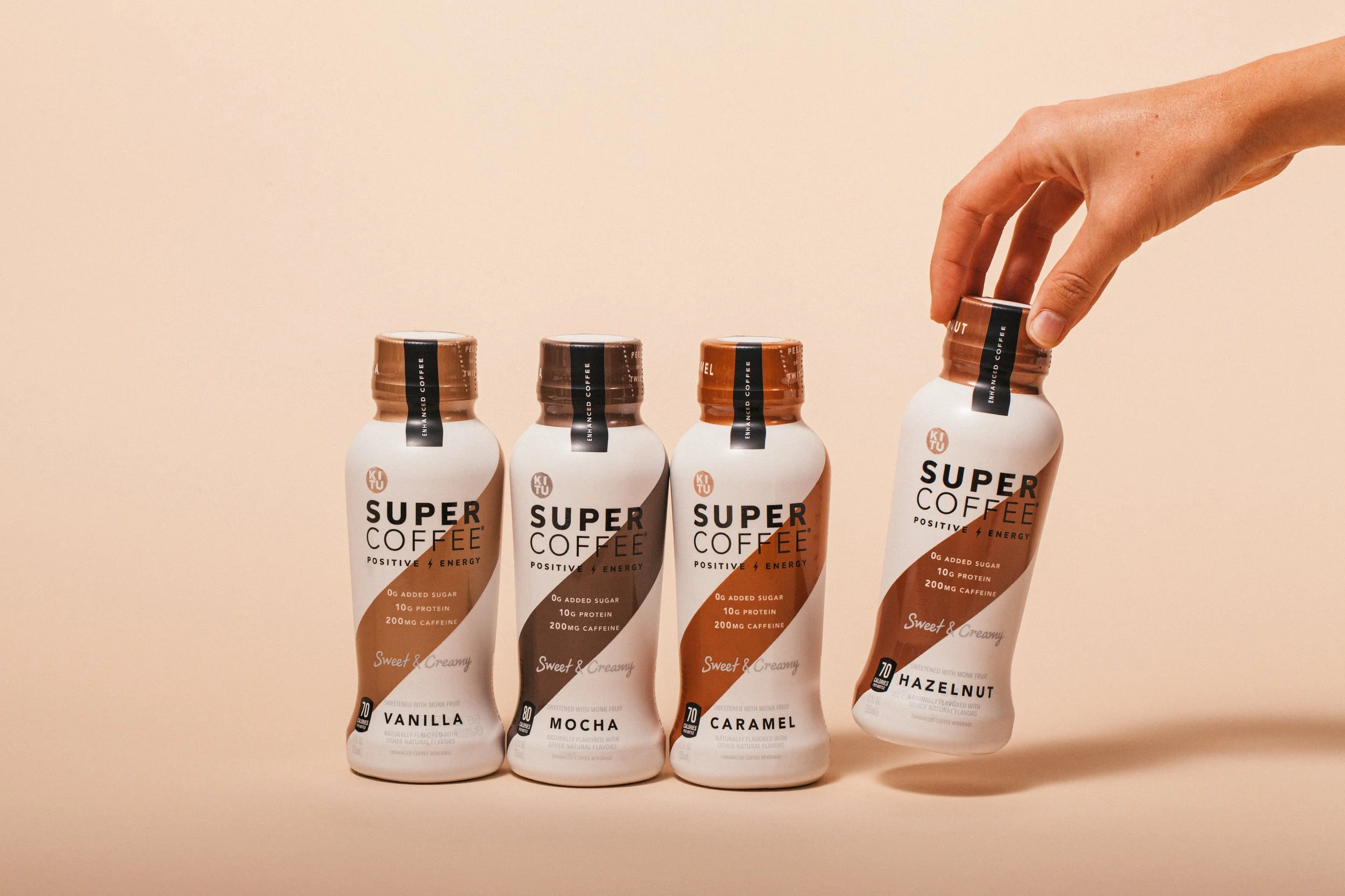 Four bottles of Super Coffee drinks labeled vanilla, mocha, caramel, and hazelnut, with a hand reaching to pick up the hazelnut flavor, all against a beige background.