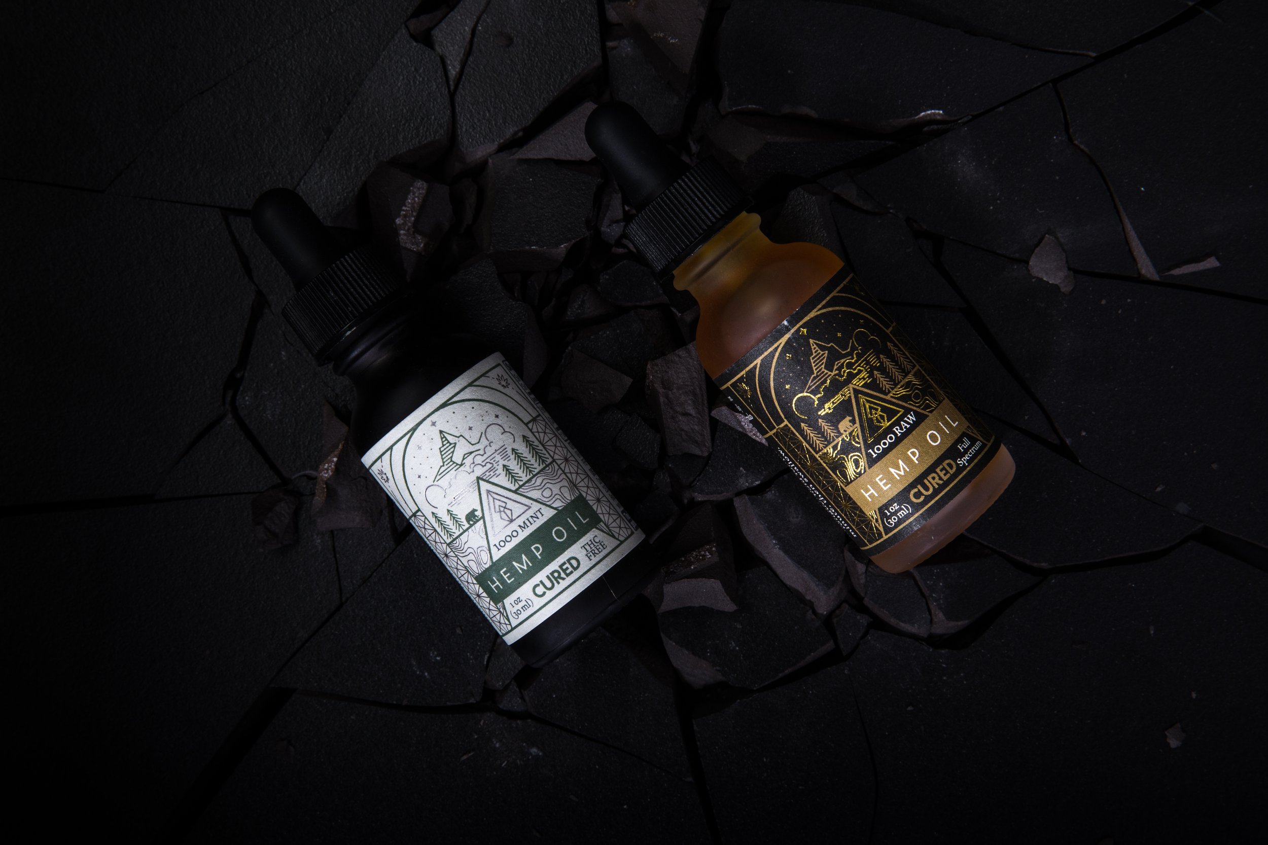Two bottles of hemp oil with dropper caps, black and gold packaging, resting on shattered black rocks.
