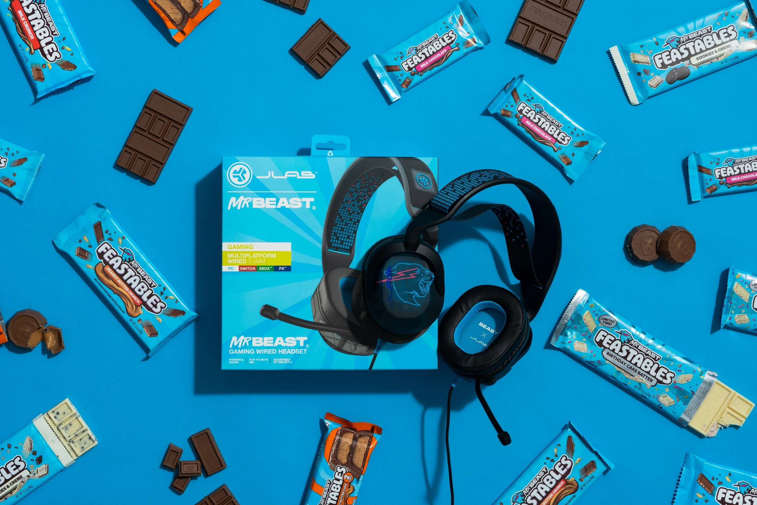 A boxed MrBeast gaming wired headset with a black and blue design, surrounded by assorted MrBeast Feastables chocolate bars and some chocolate pieces on a blue background.