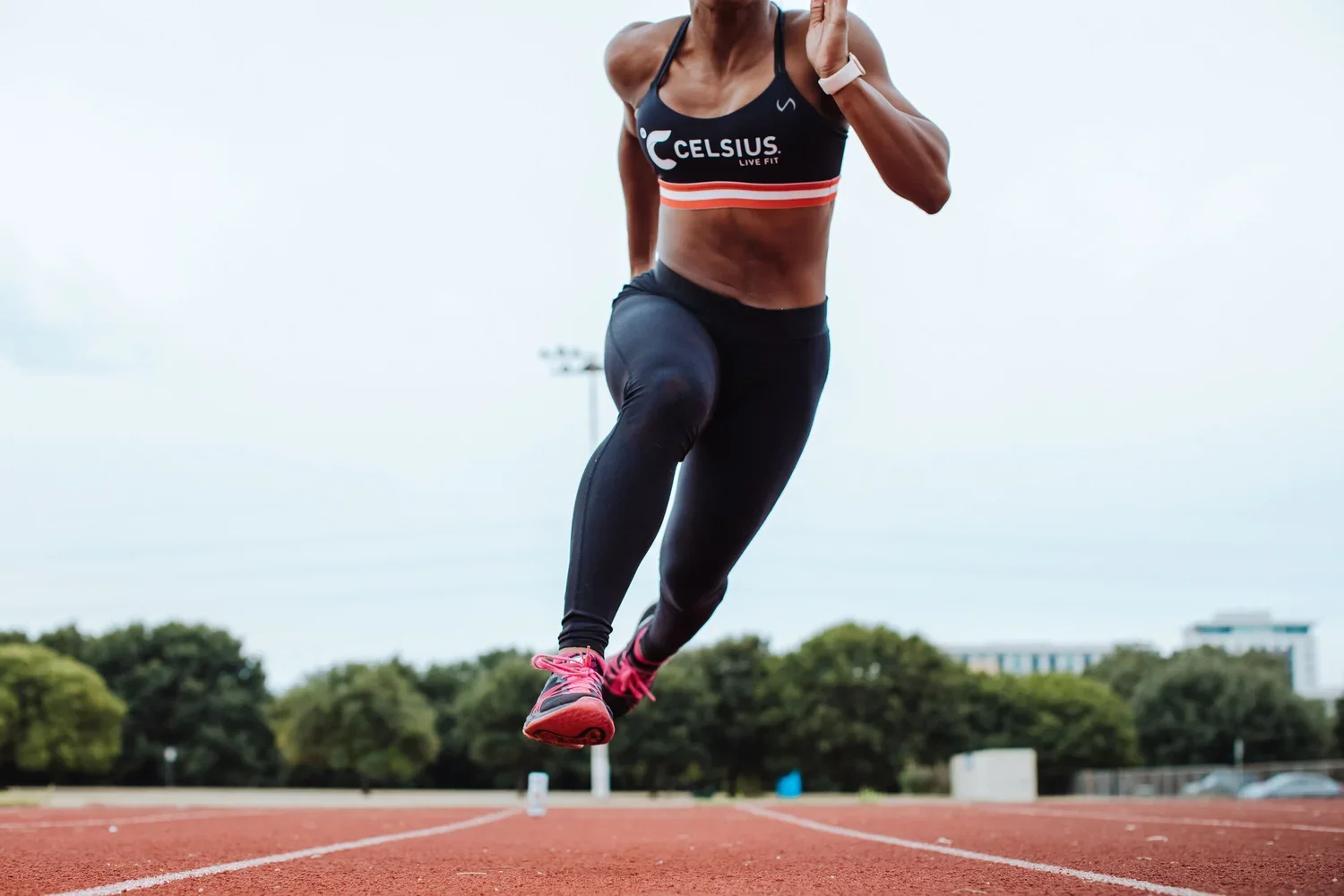 A female athlete wearing a black sports bra and black leggings is running on a track. She has pink running shoes, and her hair and face are not visible. The background shows trees and a cloudy sky.