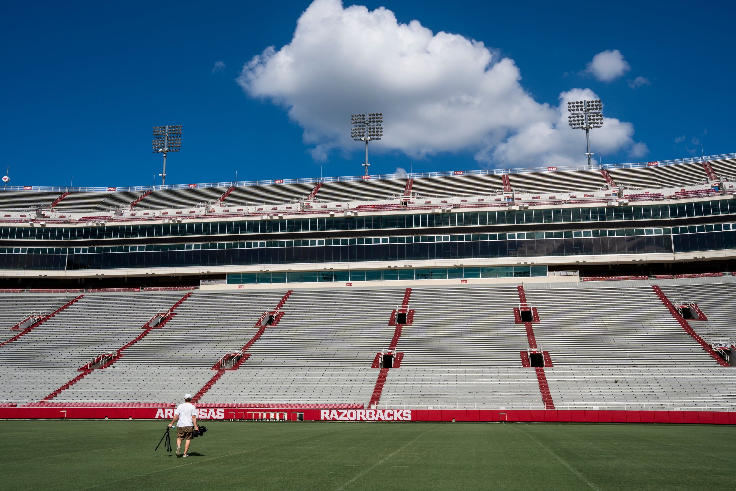 An empty football stadium with the field in the foreground, a person walking on the grass carrying a tripod, and seating stands with the Arkansas Razorbacks logo on the field barrier, under a bright blue sky with some clouds.