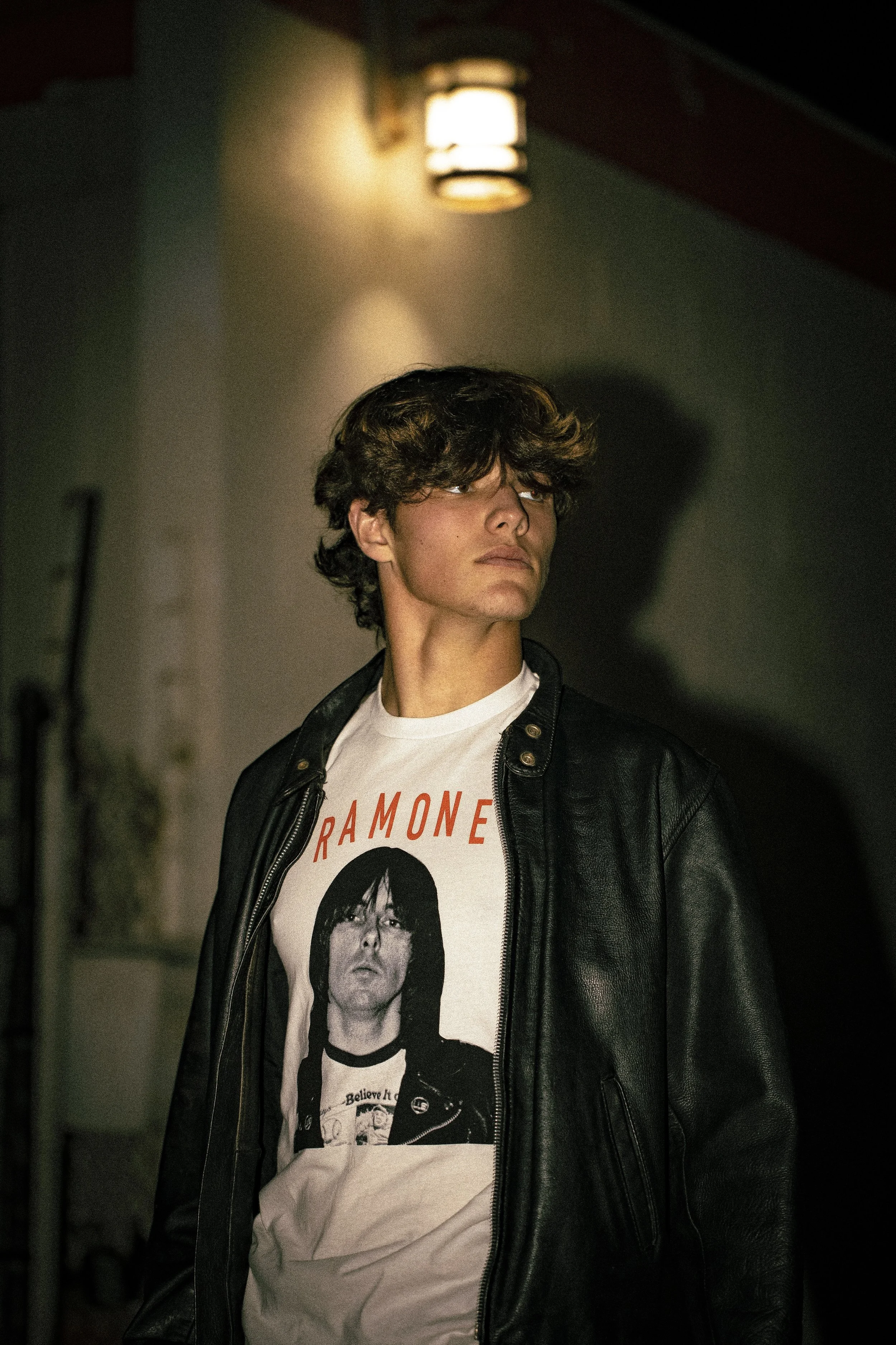 A young man with dark, wavy hair stands in a dimly lit alleyway, wearing a black leather jacket over a Ramones t-shirt with a black-and-white image of a band member on it.
