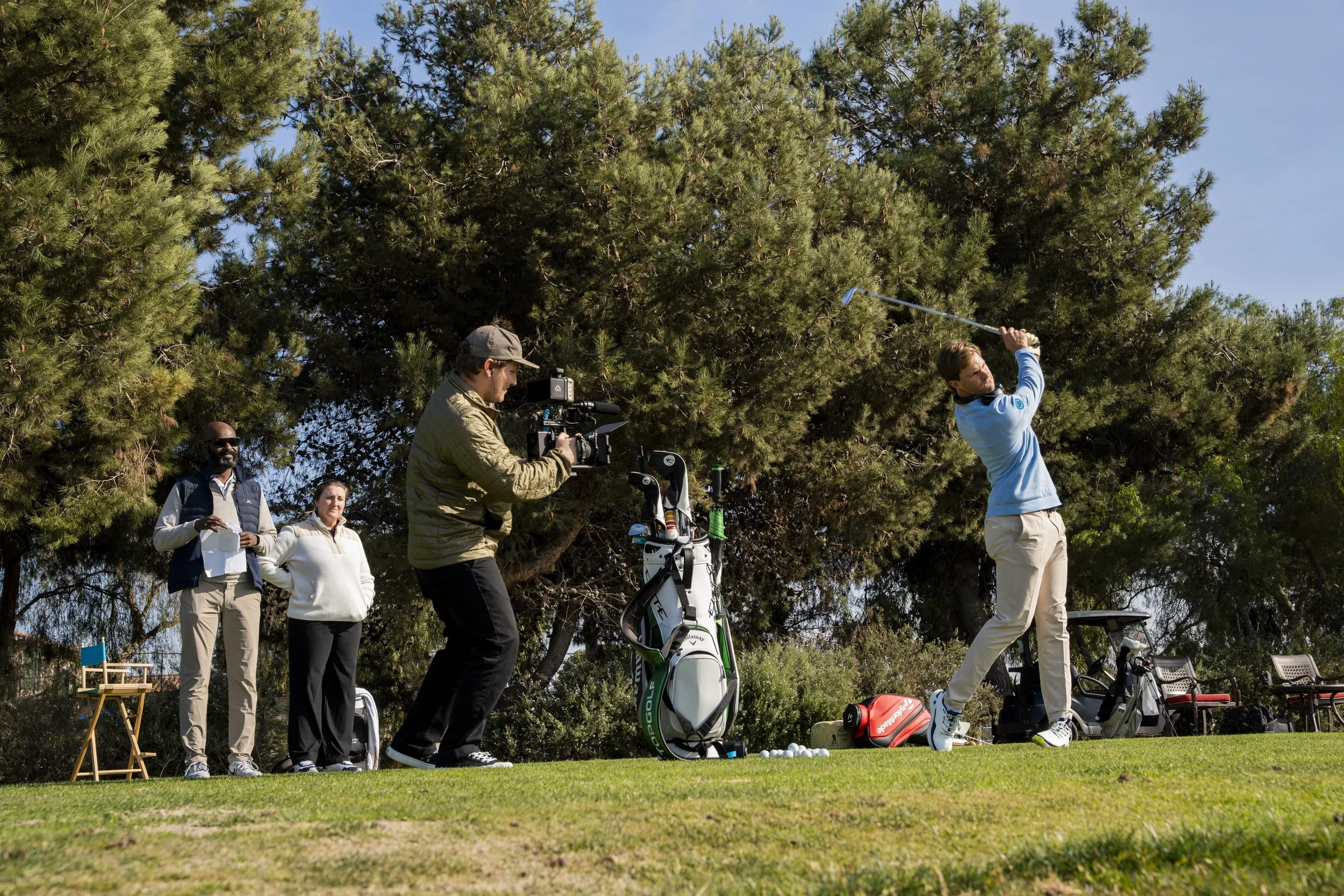 A man in a blue sweater taking a golf swing at a golf course with three people observing and a cameraman filming.