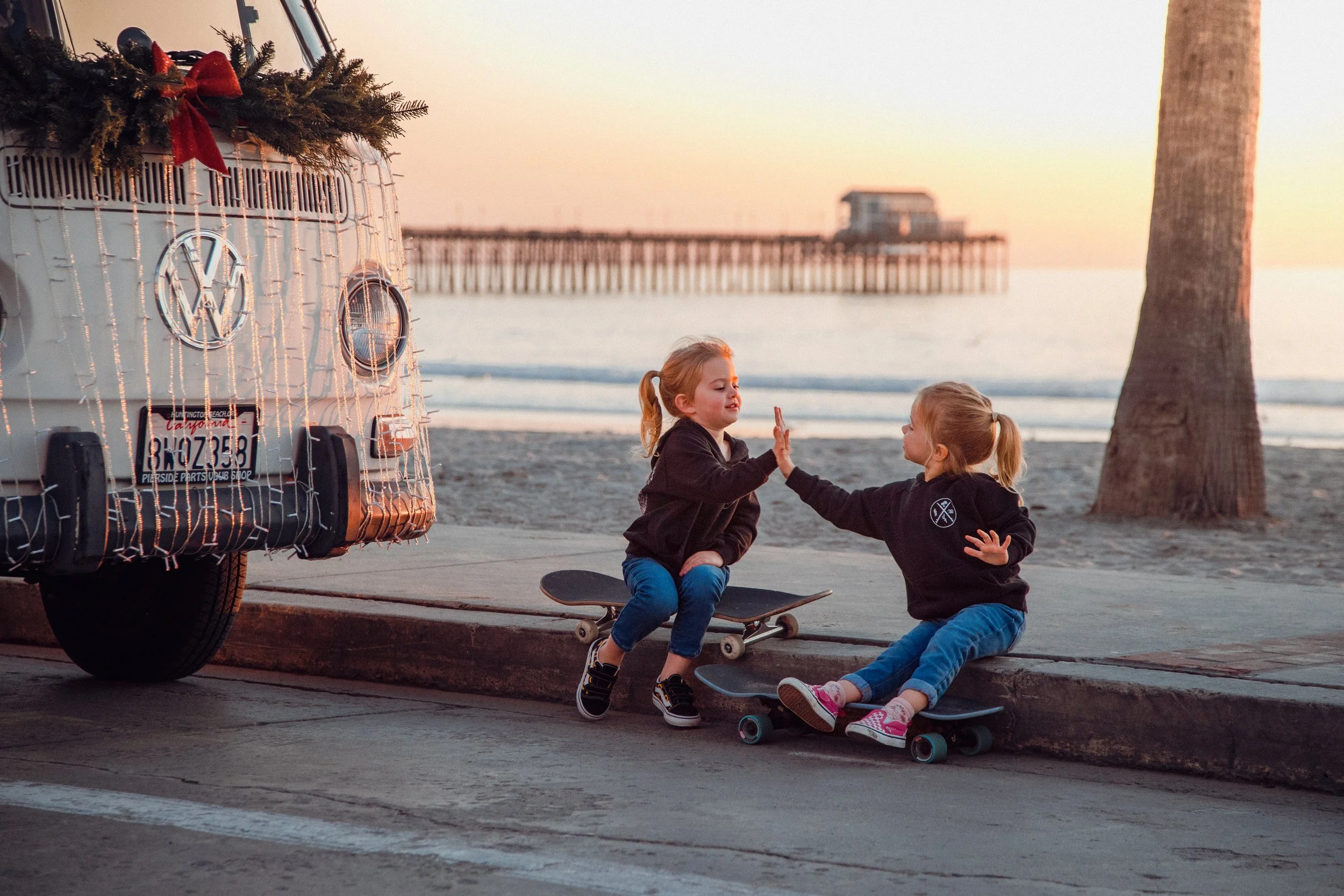Two young girls sitting on skateboards at the beach, giving each other a high five at sunset, with a decorated Volkswagen van and a pier in the background.