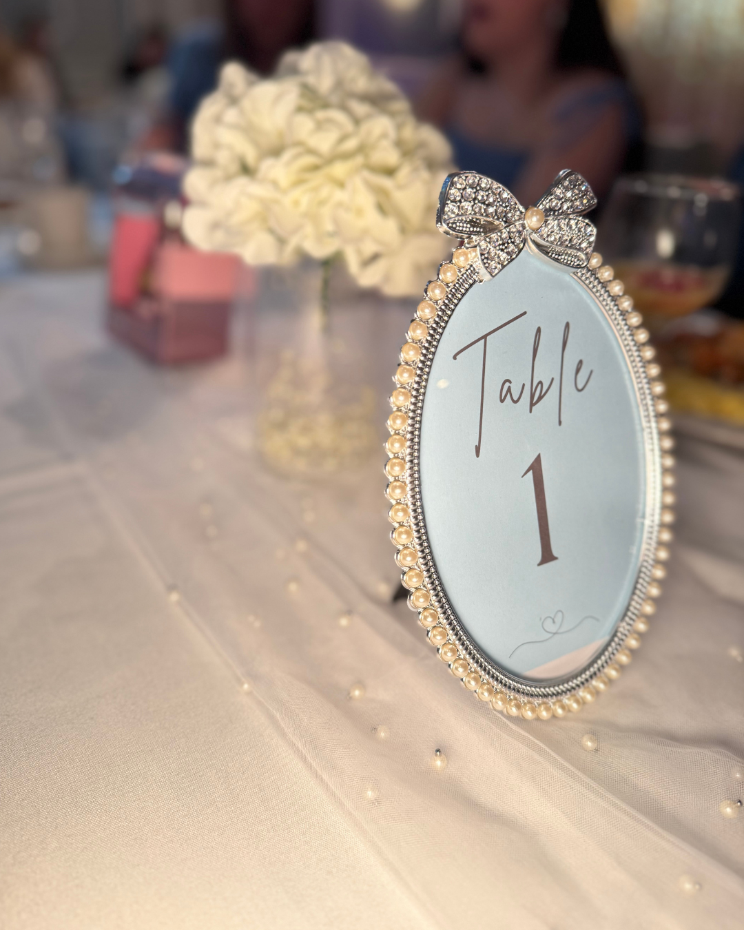 Table number sign with a pearl and rhinestone bow, labeled 'Table 1' on a decorated table with flowers and guests in the background.