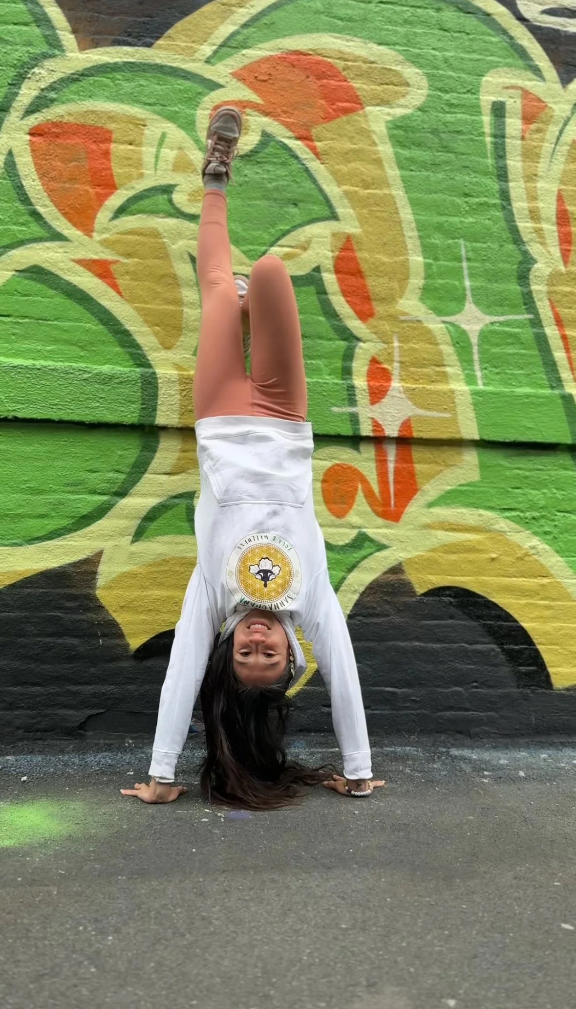 A woman performing a handstand against a colorful graffiti wall, wearing a white hoodie, beige pants, and sneakers, with her long dark hair hanging down.