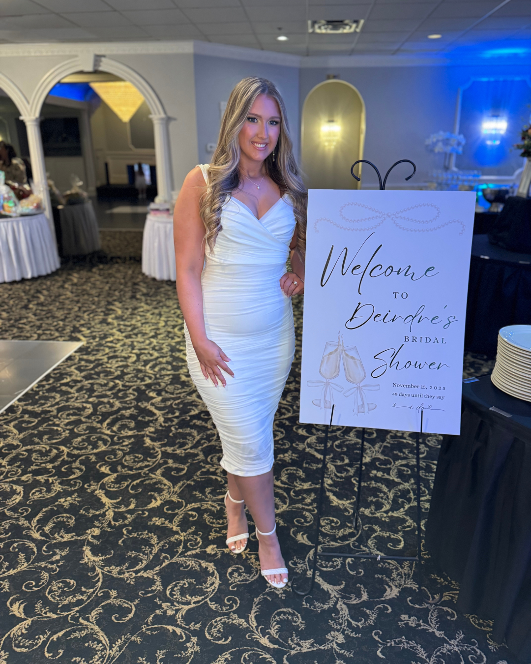 A woman in a white dress standing next to a welcome sign for a bridal shower event, in an elegantly decorated venue.